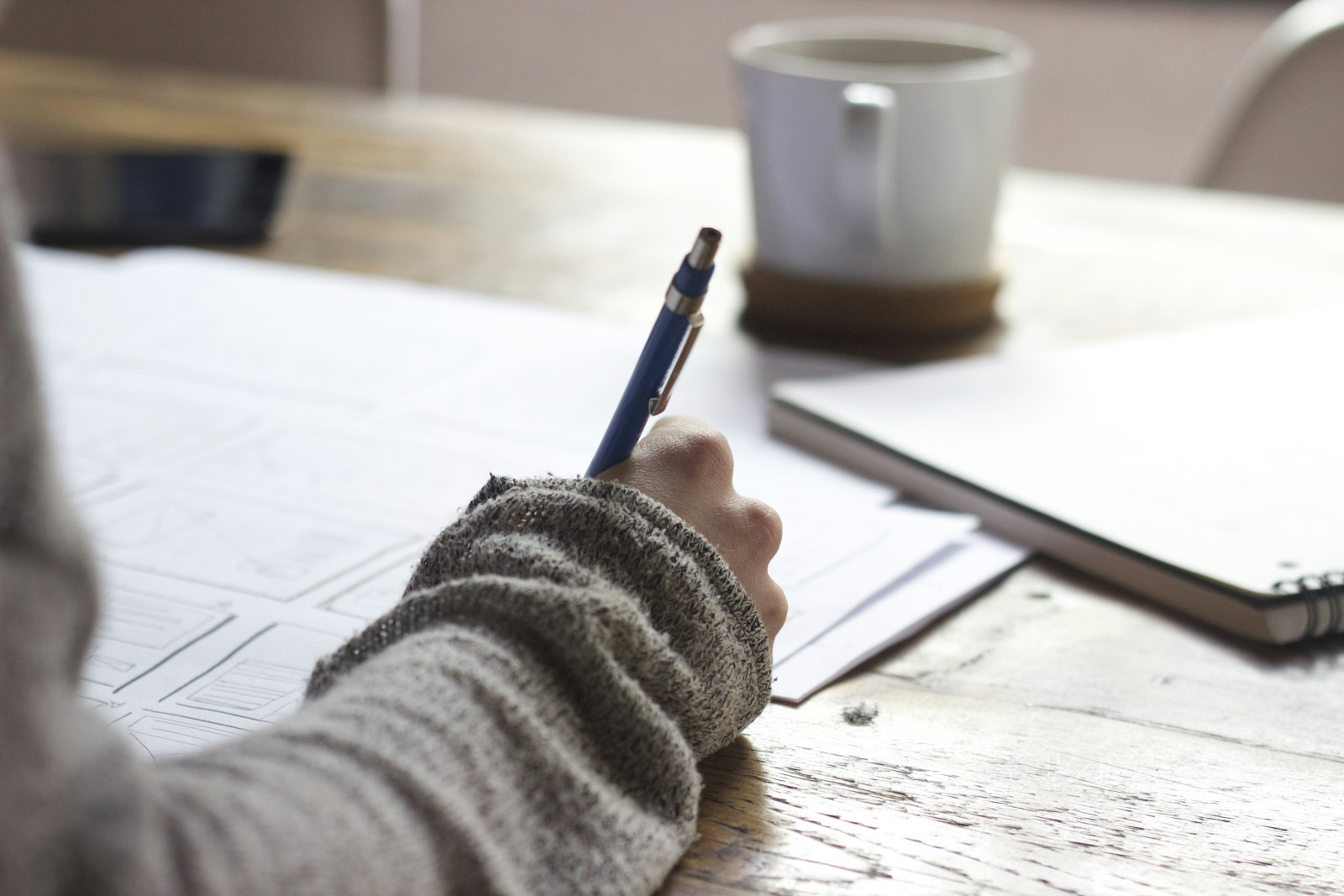 A hand writing a list in a notebook next to a cup of coffee